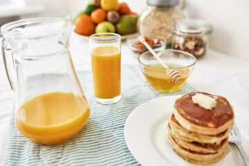 A jug and a glass of orange juice, honey in a bowl, a stack of pancakes and various fruits served for breakfast on the table