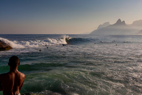 Man watching surfers surfing at sunrise in Rio de Janeiro, Brazil. Urban buildings under the sun far away and nice ocean view. Travel Destination holiday vacation new normal reopening concept.
