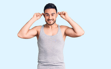 Young handsome man wearing swimwear and sleeveless t-shirt smiling pulling ears with fingers, funny...