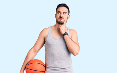 Young handsome man holding basketball ball serious face thinking about question with hand on chin, thoughtful about confusing idea