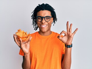 Young african american man holding nachos potato chips doing ok sign with fingers, smiling friendly...