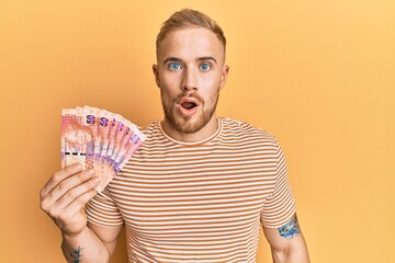 Young caucasian man holding south african 50 rand banknotes scared and amazed with open mouth for surprise, disbelief face