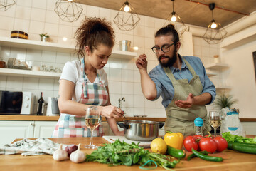 Italian man adding pepper, spice to the soup while woman stirring it with a spoon. Couple preparing a meal together in the kitchen. Cooking at home, Italian cuisine