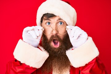 Handsome young red head man with long beard wearing santa claus costume and glasses in shock face,...