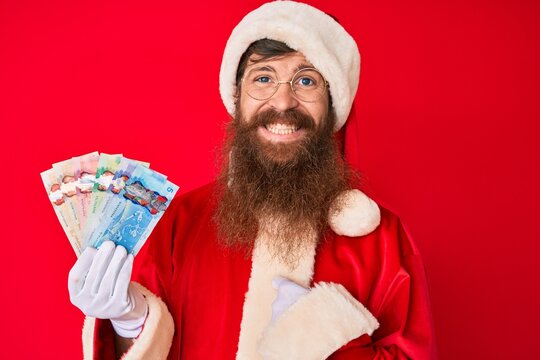 Handsome young red head man with long beard wearing santa claus costume holding canadian dollars pointing finger to one self smiling happy and proud