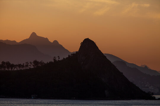 Beautiful distant view of a pointy mount silhouette at sunset against orange sky; Picture taken from Niteroi, Rio de Janeiro, Brazil. Detail of the sea and some vegetation. Copy space for text.