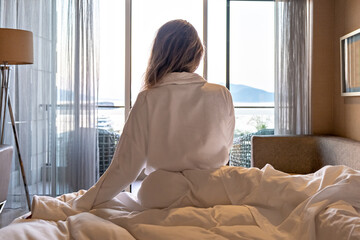back view of woman in bathrobe sitting on bed morning looking out the window and resting hotel room with sea view