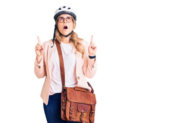 Young caucasian woman wearing bike helmet and leather bag amazed and surprised looking up and pointing with fingers and raised arms.