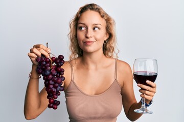 Beautiful caucasian woman holding branch of fresh grapes and red wine smiling looking to the side...
