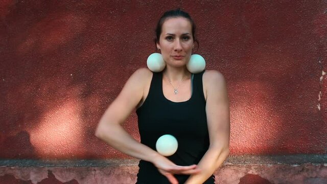 A professional juggler juggles with white balls. A professional circus actress juggles with white balls on the street. An attractive female juggler trains her juggling skills.