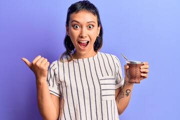 Young woman drinking mate infusion pointing thumb up to the side smiling happy with open mouth