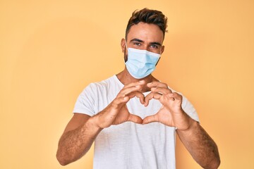 Young hispanic man wearing medical mask smiling in love doing heart symbol shape with hands....
