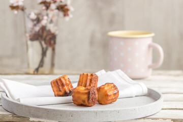 Selective focus to canelé cake - a small french pastry flavored with rum and vanilla on plate, blurred mug and flowered sprigs in the background on the wooden table.