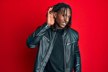African american man with braids wearing black leather jacket smiling with hand over ear listening and hearing to rumor or gossip. deafness concept.