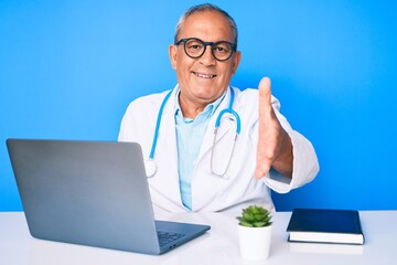 Senior handsome man with gray hair wearing doctor uniform working using computer laptop smiling friendly offering handshake as greeting and welcoming. successful business.
