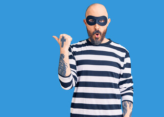 Naklejka premium Young handsome man wearing burglar mask surprised pointing with hand finger to the side, open mouth amazed expression.