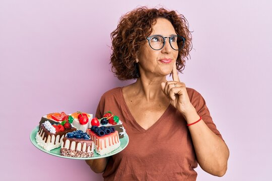 Beautiful Middle Age Mature Woman Holding Cake Slices Serious Face Thinking About Question With Hand On Chin, Thoughtful About Confusing Idea