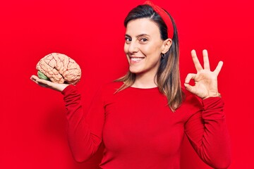 Young beautiful woman holding brain doing ok sign with fingers, smiling friendly gesturing...