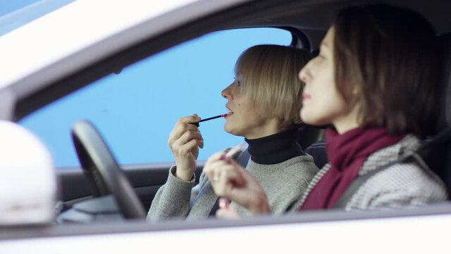 Brunette and blonde women in casual clothes are travelling. Brunette woman is driving car with greenscreen background. She is sitting in her car with her friend. They are looking at the mirror and