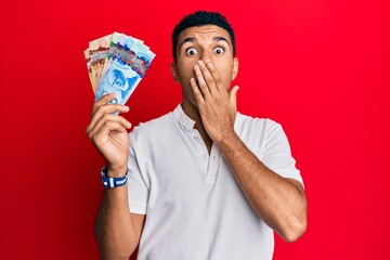 Young arab man holding canadian dollars covering mouth with hand, shocked and afraid for mistake. surprised expression