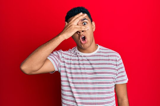 Young arab man wearing casual clothes peeking in shock covering face and eyes with hand, looking through fingers afraid
