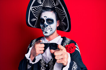 Young man wearing mexican day of the dead costume over red pointing fingers to camera with happy and funny face. good energy and vibes.