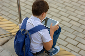 A brunette schoolboy in a white shirt and a blue backpack sits on the stairs and plays with a tablet. Back view