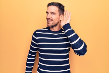 Young handsome man wearing casual striped sweater standing over isolated yellow background smiling with hand over ear listening and hearing to rumor or gossip. Deafness concept.