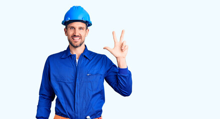 Young handsome man wearing worker uniform and hardhat showing and pointing up with fingers number three while smiling confident and happy.