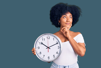 Young african american girl holding big clock serious face thinking about question with hand on...