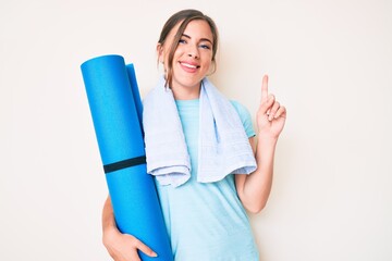Beautiful young caucasian woman holding yoga mat surprised with an idea or question pointing finger with happy face, number one