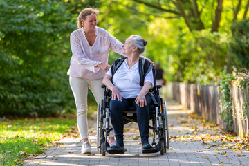 a young woman pushes an old lady in a wheelchair through a park