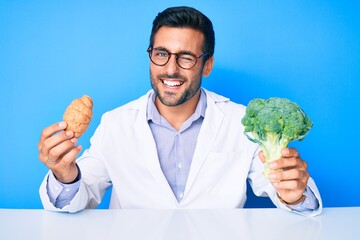 Young hispanic man as nutritionist doctor holding croissant and broccoli winking looking at the camera with sexy expression, cheerful and happy face. - obrazy, fototapety, plakaty