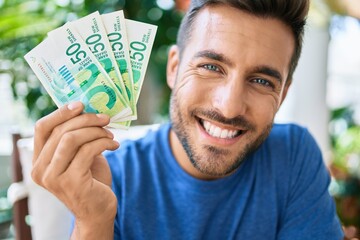 Young hispanic man smiling happy holding Israeli shekels banknotes at the terrace.