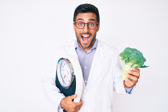 Young hispanic man as nutritionist doctor holding weighing machine and broccoli celebrating crazy and amazed for success with open eyes screaming excited.