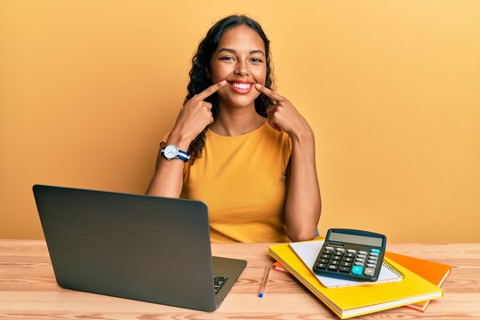 Young african american girl working at the office with laptop and calculator smiling with open mouth, fingers pointing and forcing cheerful smile