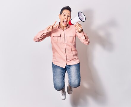 Young beautiful caucasian woman screaming using megaphone. Jumping doing victory sign over isolated white background