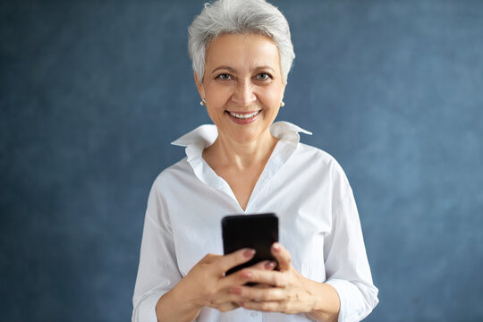 Happy Energetic 50 Year Old Woman Marketing Expert In White Formal Shirt Posing In Studio With Smart Phone, Typing Post For Social Media Account, Smiling. Electronic Gadgets And Occupation