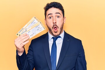 Young hispanic man wearing suit holding boarding pass scared and amazed with open mouth for surprise, disbelief face