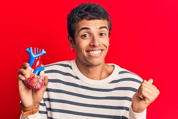 Young african amercian man wearing holding heart screaming proud, celebrating victory and success very excited with raised arm