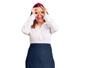 Young latin woman wearing waitress apron doing ok gesture like binoculars sticking tongue out, eyes looking through fingers. crazy expression.