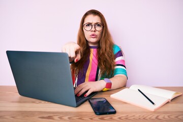 Young redhead woman working at the office with laptop pointing with finger to the camera and to you, confident gesture looking serious