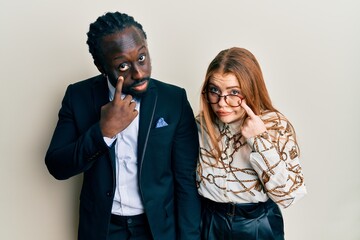 Young interracial couple wearing business and elegant clothes pointing to the eye watching you gesture, suspicious expression