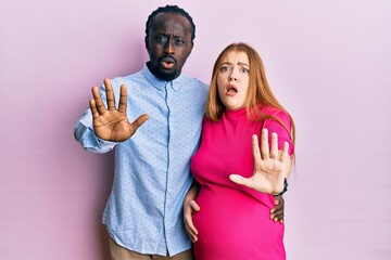 Young interracial couple expecting a baby, touching pregnant belly doing stop gesture with hands palms, angry and frustration expression