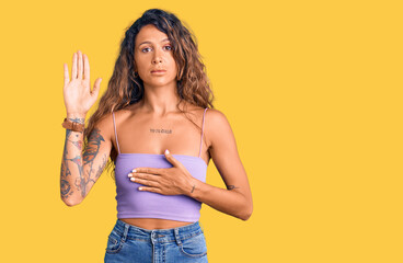 Young hispanic woman with tattoo wearing casual clothes swearing with hand on chest and open palm,...
