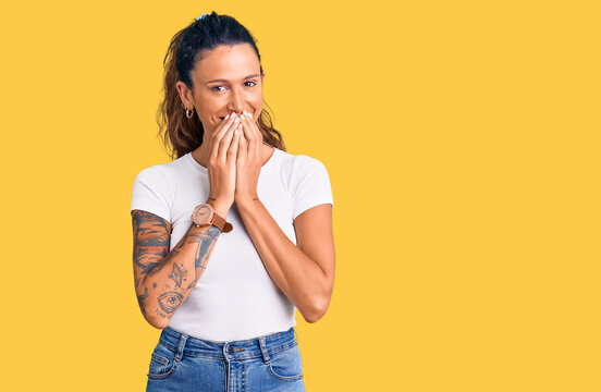 Young hispanic woman with tattoo wearing casual white tshirt laughing and embarrassed giggle covering mouth with hands, gossip and scandal concept