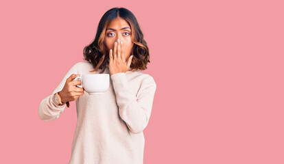 Young beautiful mixed race woman holding a cup of coffee covering mouth with hand, shocked and afraid for mistake. surprised expression