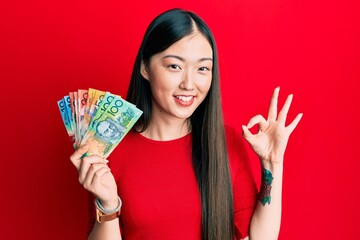 Young chinese woman holding australian dollars doing ok sign with fingers, smiling friendly gesturing excellent symbol