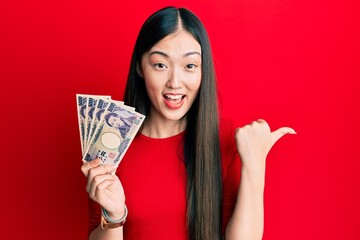 Young chinese woman holding japanese yen banknotes pointing thumb up to the side smiling happy with open mouth