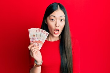 Young chinese woman holding 10 colombian pesos banknotes scared and amazed with open mouth for surprise, disbelief face